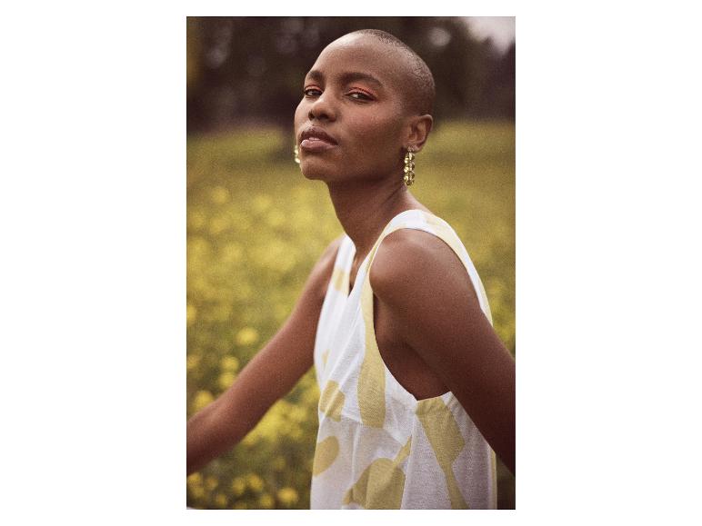 Model in mouwloze jurk met abstract print in een veld met gele bloemen.