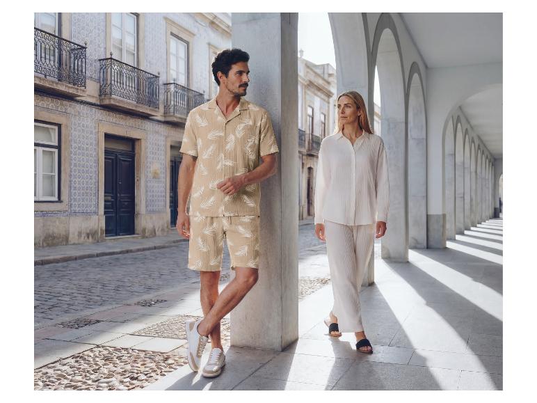 Homme en ensemble chemise et short à motifs de feuilles, femme en chemise et pantalon blancs, dans une rue à arcades.