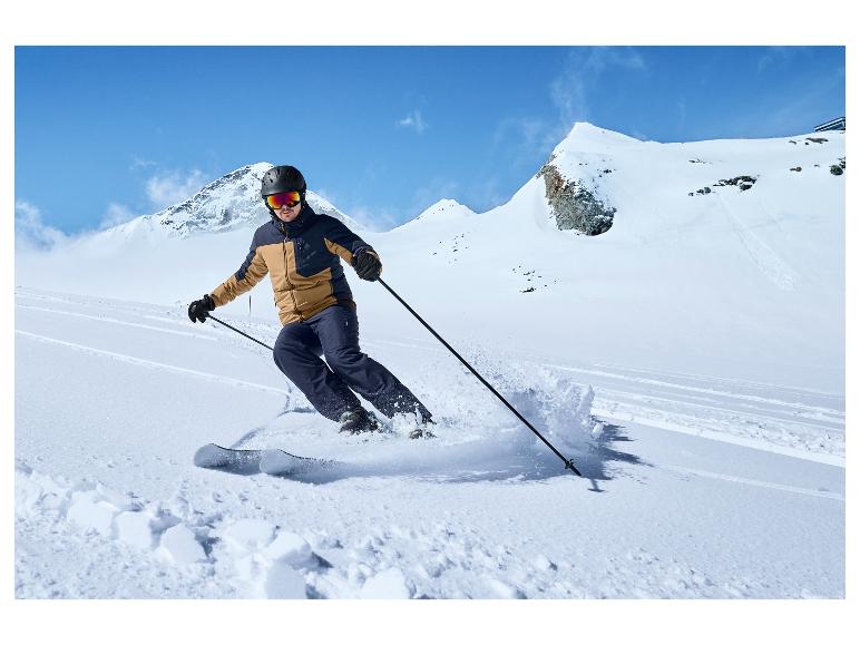 Un skieur avec un pantalon bleu et une veste marron descend une pente à ski.