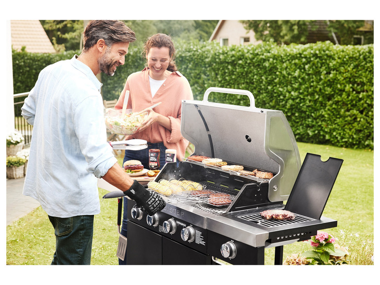 Een stel kookt hamburgers, worstjes en maïs op een zwarte gasbarbecue.