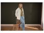 Vrouw in groene trui, wit jack en blauwe jeans, leunend tegen een houten stoel.