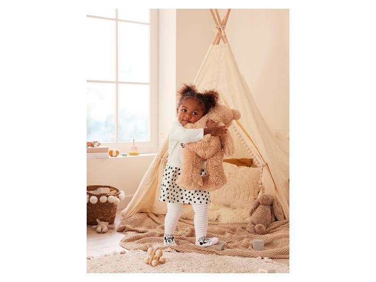 Une petite fille portant une robe et des chaussettes Mickey Mouse enroule ses bras autour d'un ours en peluche devant une tente tipi.