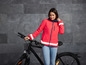 Een vrouw in een felroze jas met reflecterende strepen staat naast een zwarte fiets.