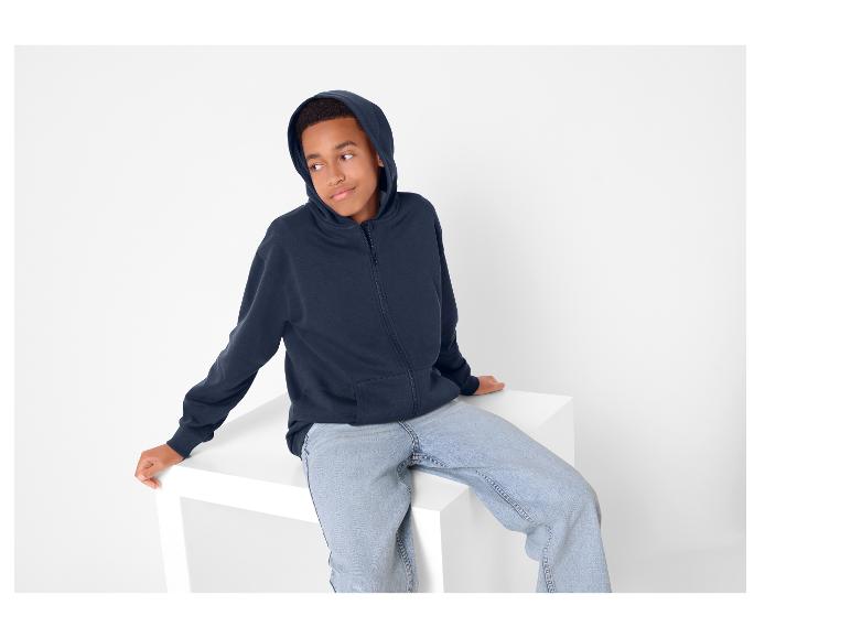Garçon en sweat à capuche zippé bleu marine et jean bleu, assis sur un cube blanc