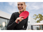 Femme souriante en pull noir tenant une raquette de tennis de table rouge à l'extérieur.