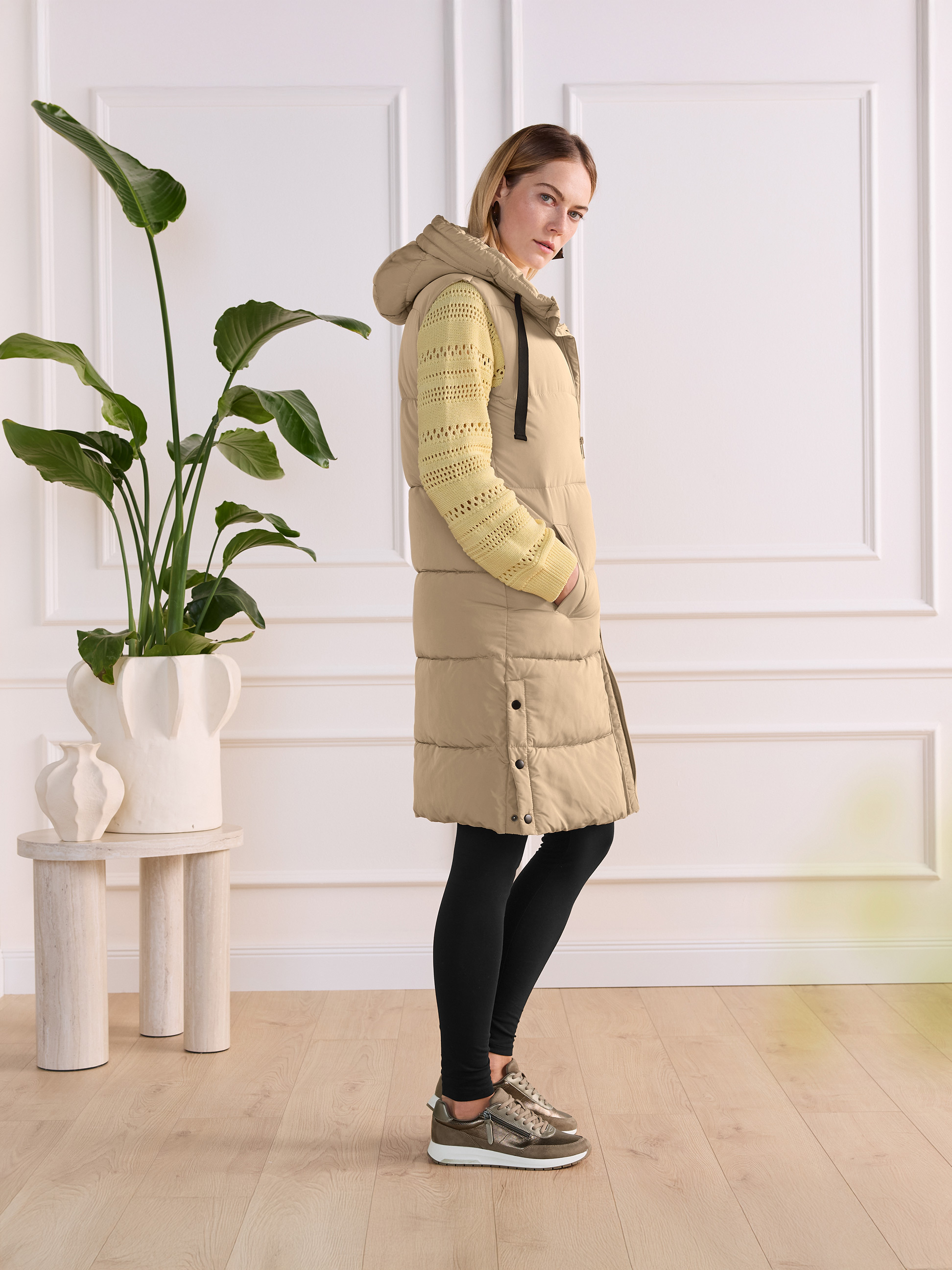 Femme en longue veste beige, pull tricoté jaune et leggings noirs avec baskets.