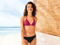 Een vrouw in een roze bikini staat voor een strand met een rieten achtergrond.