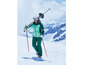 Een vrouw in een groene ski-outfit loopt met ski's op haar rug over besneeuwde berghellingen.