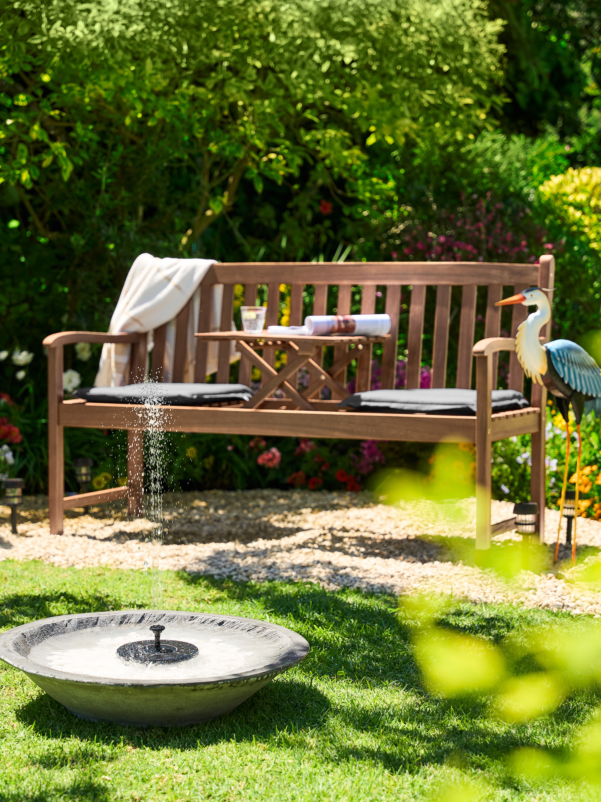 Banc de jardin en bois avec une petite table, des coussins et un héron décoratif dans un jardin ensoleillé.