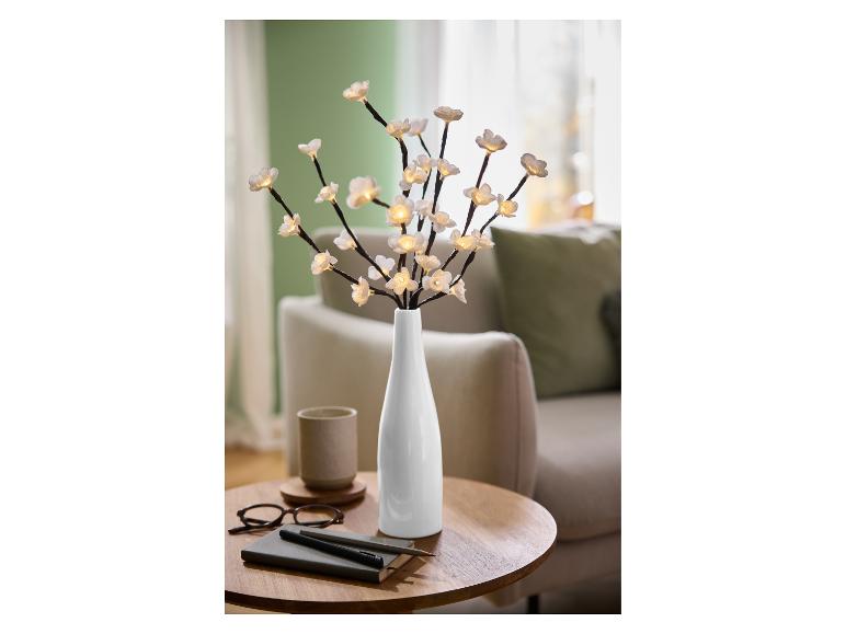 LED-lichtgevende takken met witte bloemen in een vaas, op een houten tafel met notitieboekje en bril.