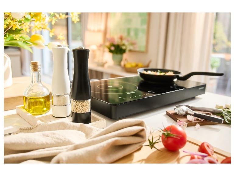 Table de cuisson à induction avec poêle, moulins à épices, huile d'olive et légumes frais sur un comptoir de cuisine.