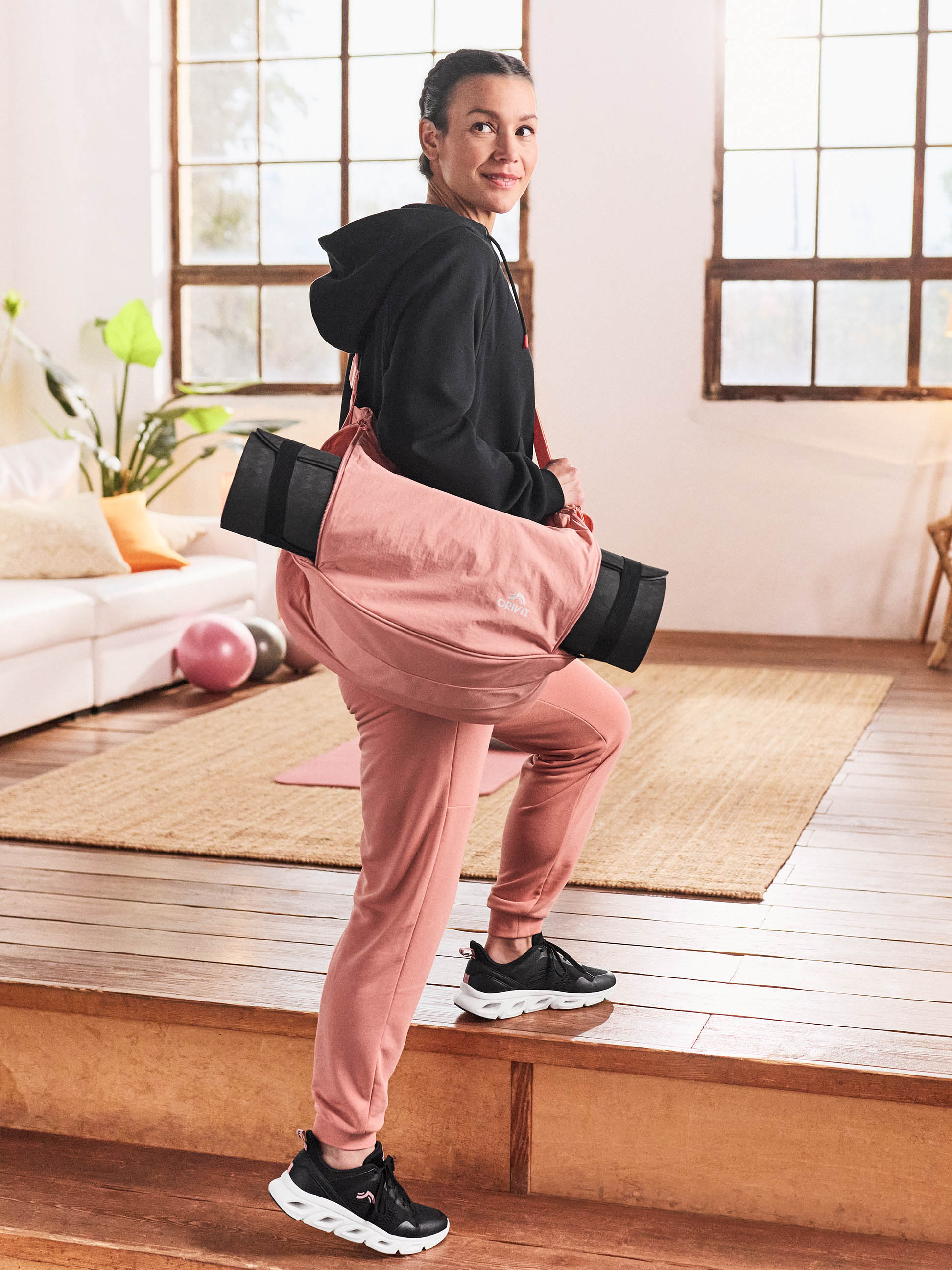 Vrouw in zwarte hoodie en roze joggingbroek met yogamat over haar schouder.
