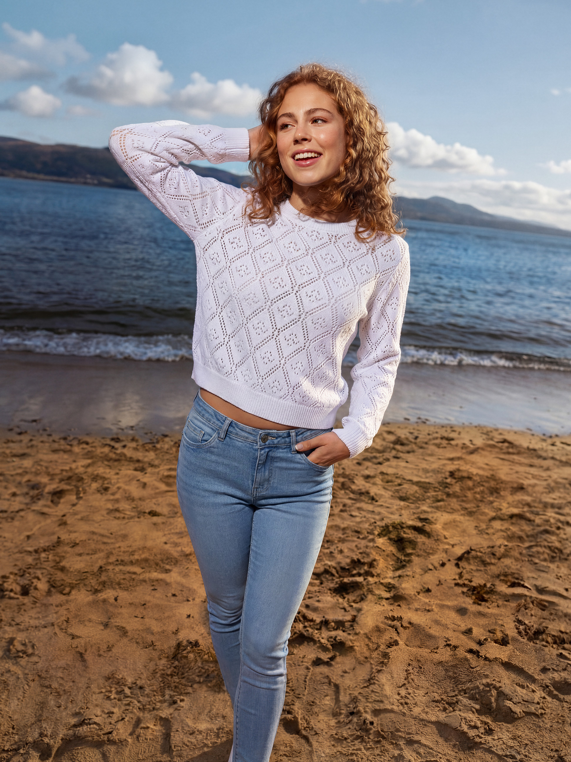 Femme en pull blanc et jean sur la plage, avec mer et montagnes en arrière-plan.