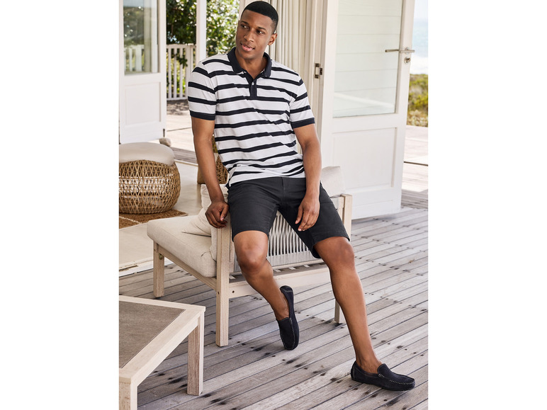 Man in een gestreepte polo, korte broek en loafers op een terras.