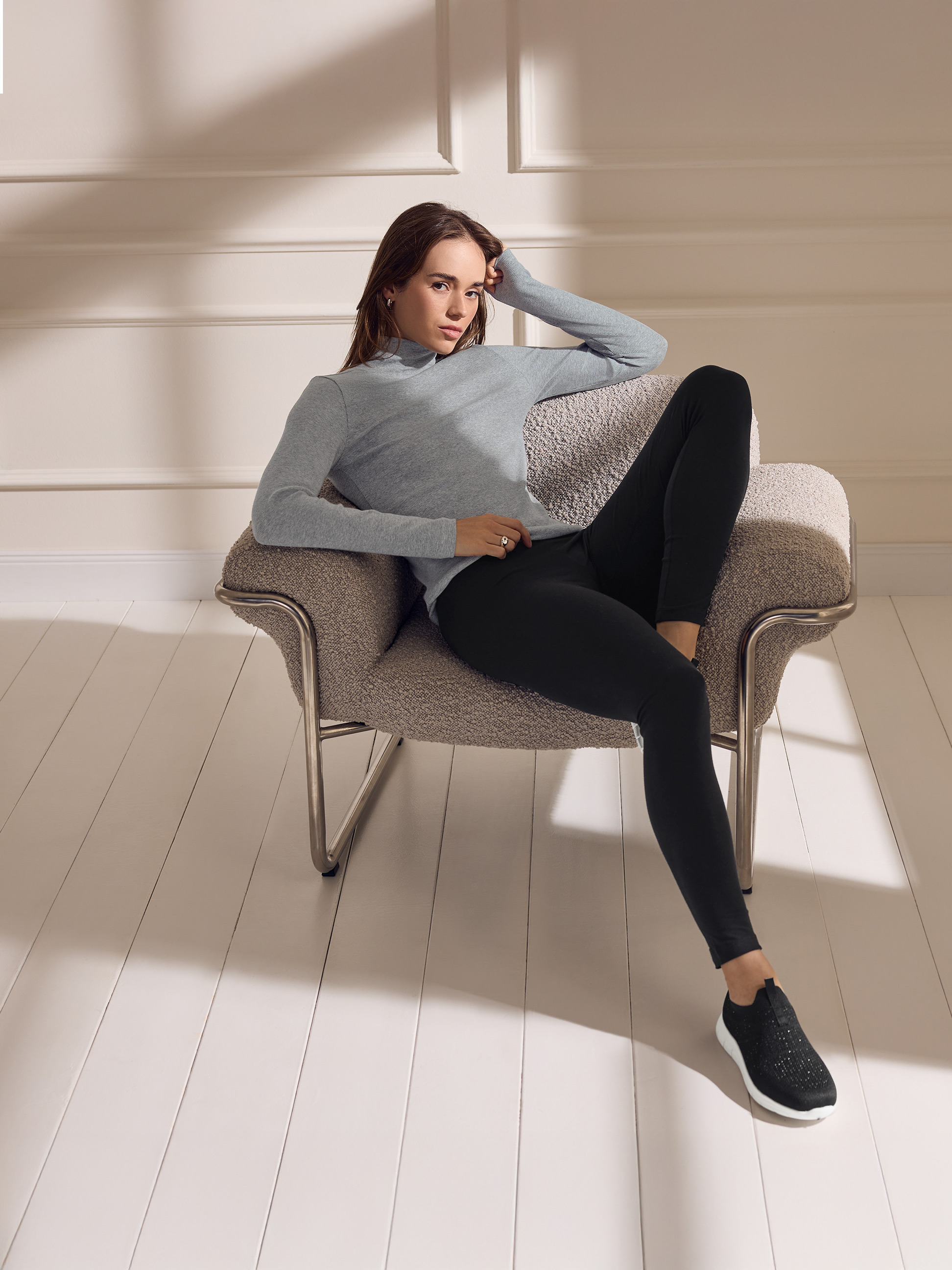 Femme en pull à col roulé gris et leggings noirs, assise sur une chaise.