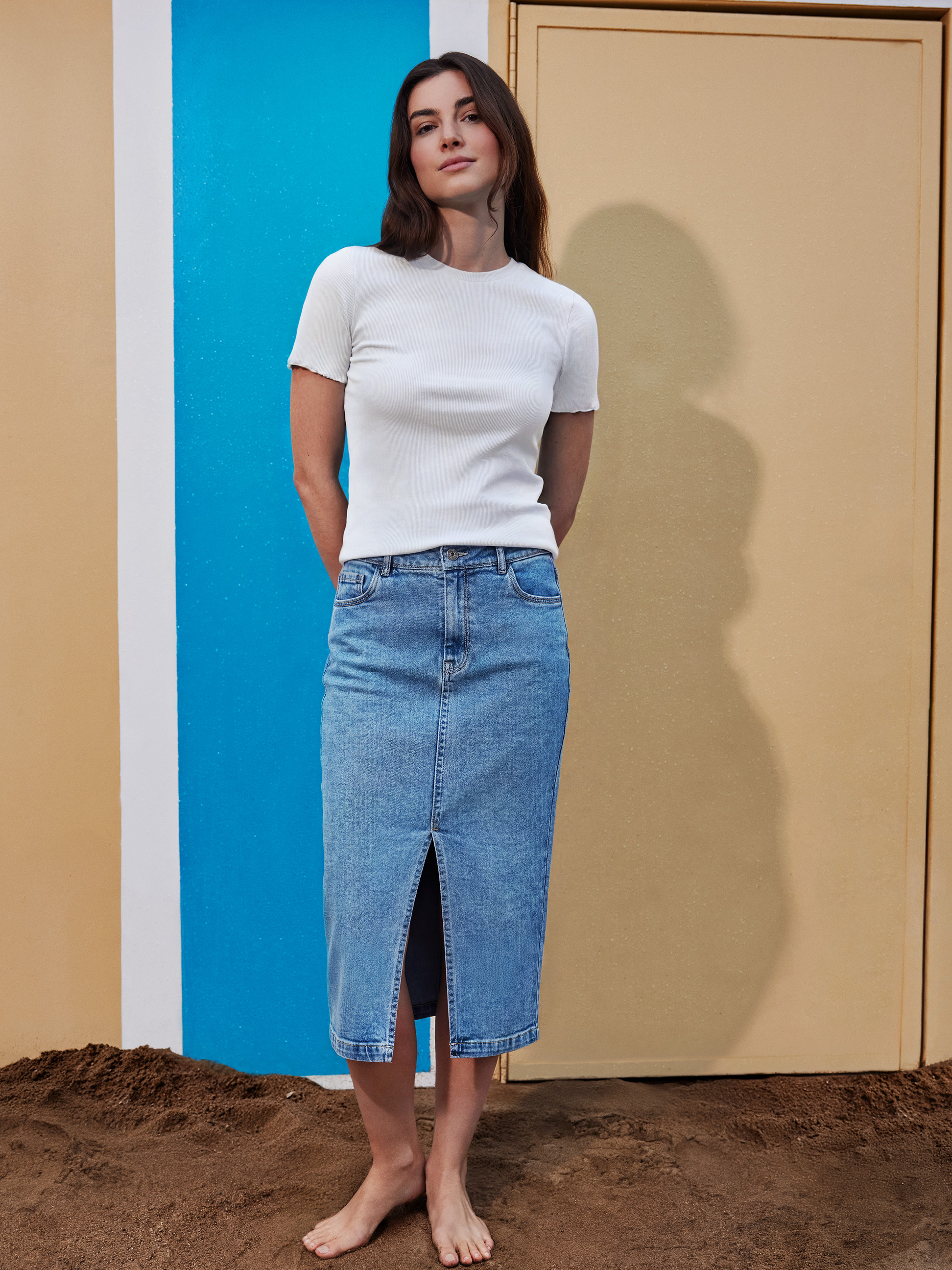 Femme en haut blanc et jupe midi en jean fendue, pieds nus sur le sable.