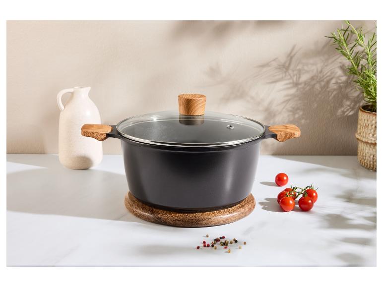 Marmite noire avec poignées en bois et couvercle en verre sur un support en bois, avec tomates et épices.