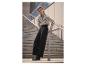 Femme en chemise à motifs noirs et blancs et pantalon noir, appuyée contre une rampe d'escalier.
