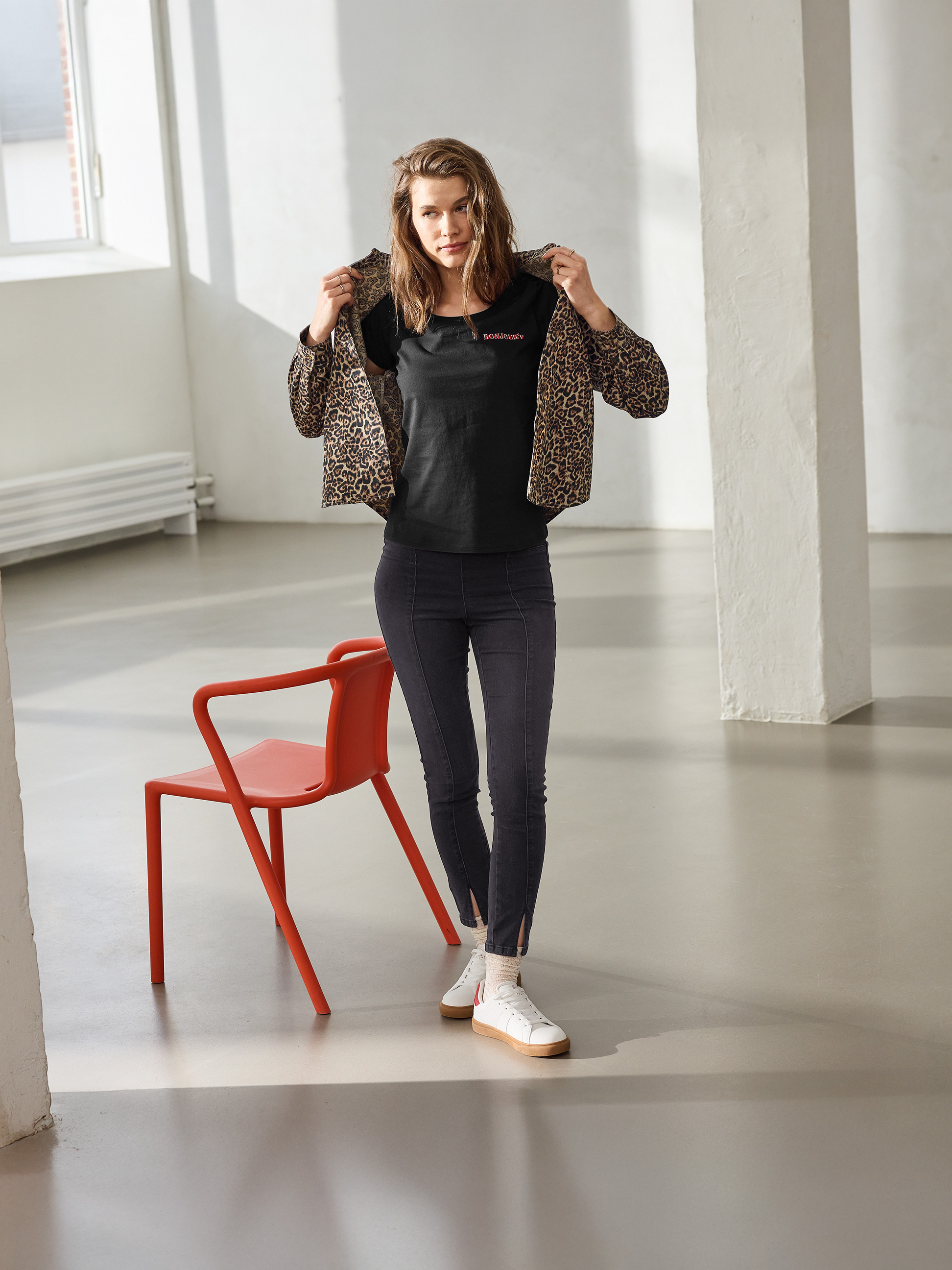 Femme en veste léopard, t-shirt noir 'BONJOUR' et jean slim noir, posant à côté d'une chaise rouge.