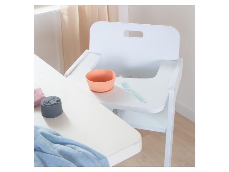 Witte kinderstoel met oranje kom en blauwe lepel, naast een tafel met een grijze houder.