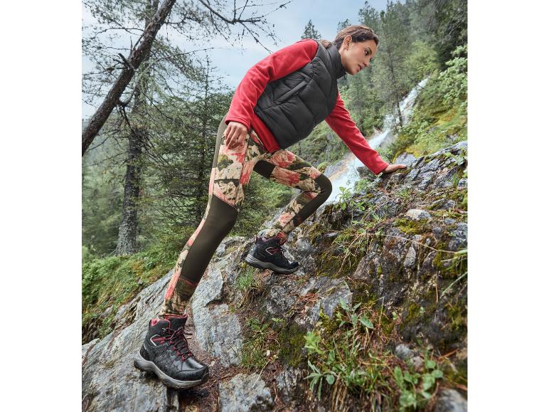 Femme en veste rouge et pantalon à imprimé floral marchant sur des rochers.