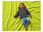 Un bébé souriant en cardigan gris et pantalon bleu, allongé sur une couverture verte.