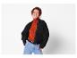 Femme rousse souriante portant une veste en velours côtelé noir, un col roulé orange et un jean.