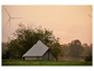 Une tente blanche dans un pré au lever du soleil, avec des éoliennes au loin.