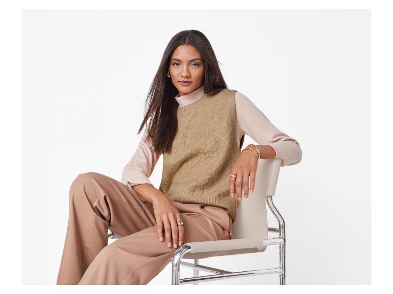 Vrouw in een beige gebreid vest en bijpassende broek, zittend op een moderne stoel.