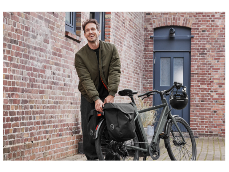 Homme souriant debout à côté d'un vélo avec des sacoches devant un mur de briques.
