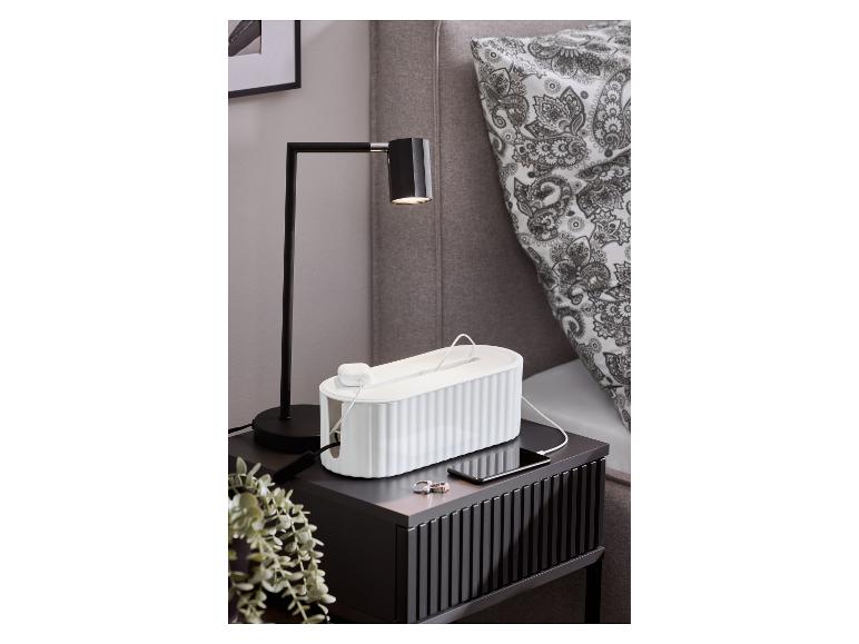 Table de chevet noire avec une lampe de table, une boîte à câbles blanche et un smartphone en charge.