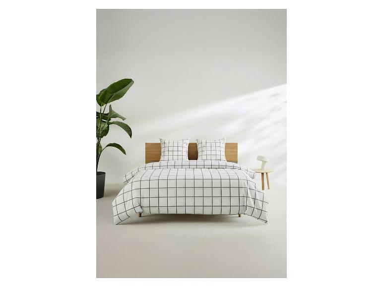 Lit avec parure de lit à carreaux, tête de lit en bois, plante et table de chevet avec lampe.