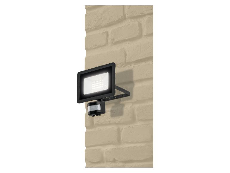 Zwarte LED-schijnwerper met bewegingssensor, gemonteerd op een beige bakstenen muur.