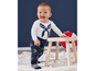 Un bébé dans un t-shirt blanc à rayures bleues et un pantalon bleu à imprimé bateaux se tient debout sur un tabouret en bois.