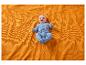 Bébé souriant en grenouillère bleue allongé sur une couverture orange texturée.