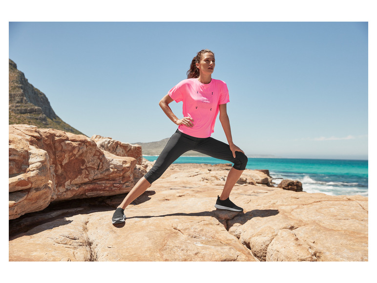 Une femme porte un t-shirt rose et un legging noir sur une côte rocheuse.