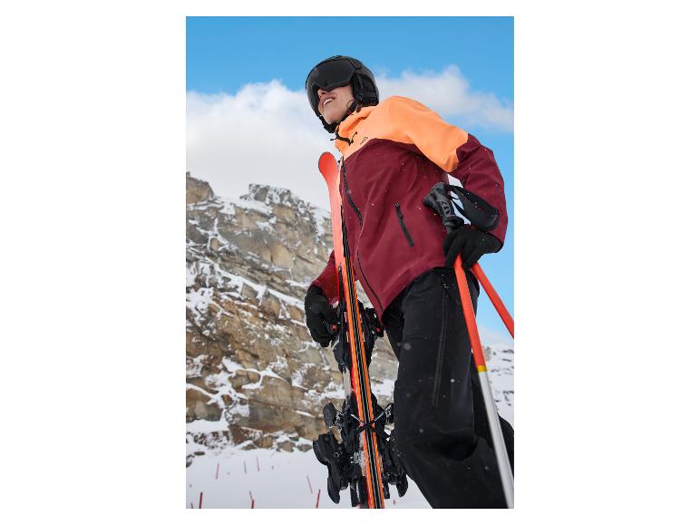 Skieur en veste orange-bordeaux, pantalon noir et casque avec lunettes, tenant skis et bâtons.