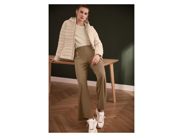 Vrouw in beige gewatteerde jas, beige T-shirt en bruine knoopbroek, leunend op een tafel.