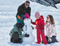 Een familie bouwt een sneeuwpop in de sneeuw, de kinderen dragen rode en groene winterkleding.
