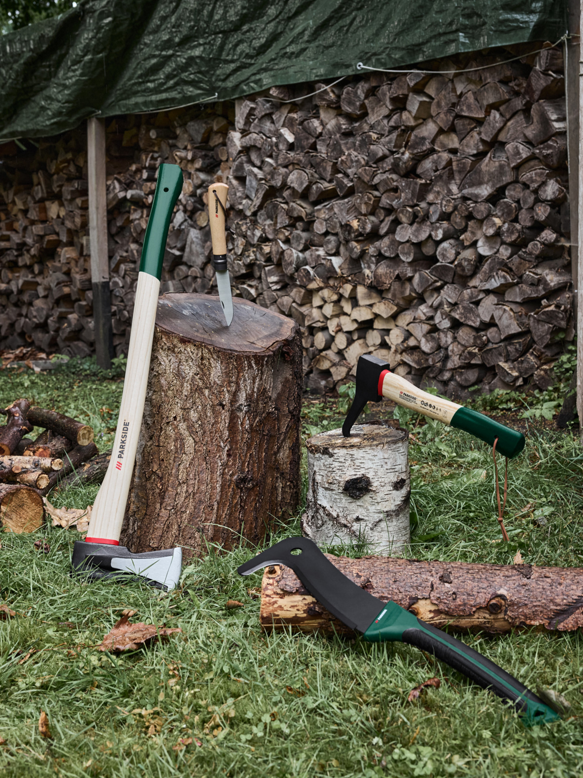 Bijlen, een mes en een machete op gras met gestapeld brandhout op de achtergrond.