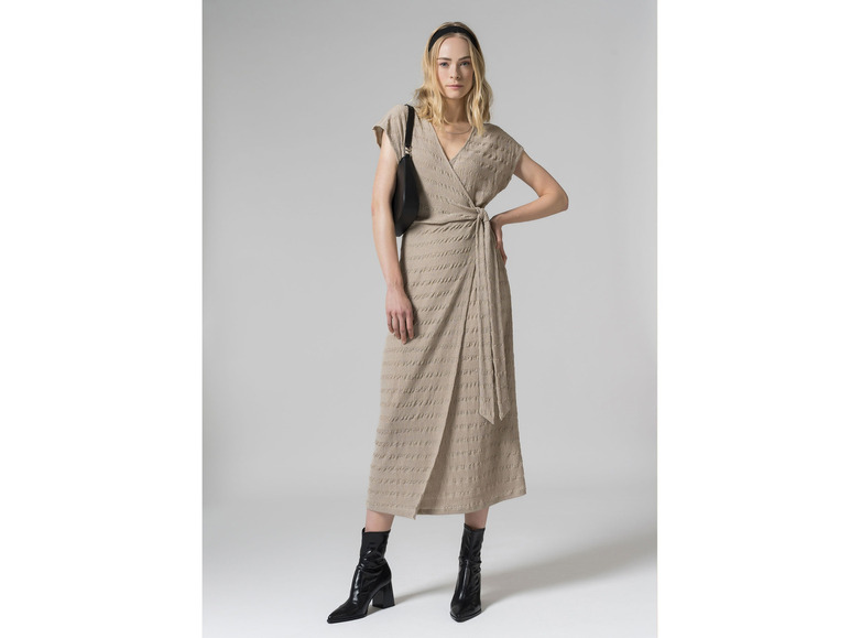 Een vrouw in een beige jurk, zwarte laarzen en een zwarte tas.