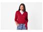Een vrouw in een rood wikkelvest en jeans met haar handen in haar zakken.