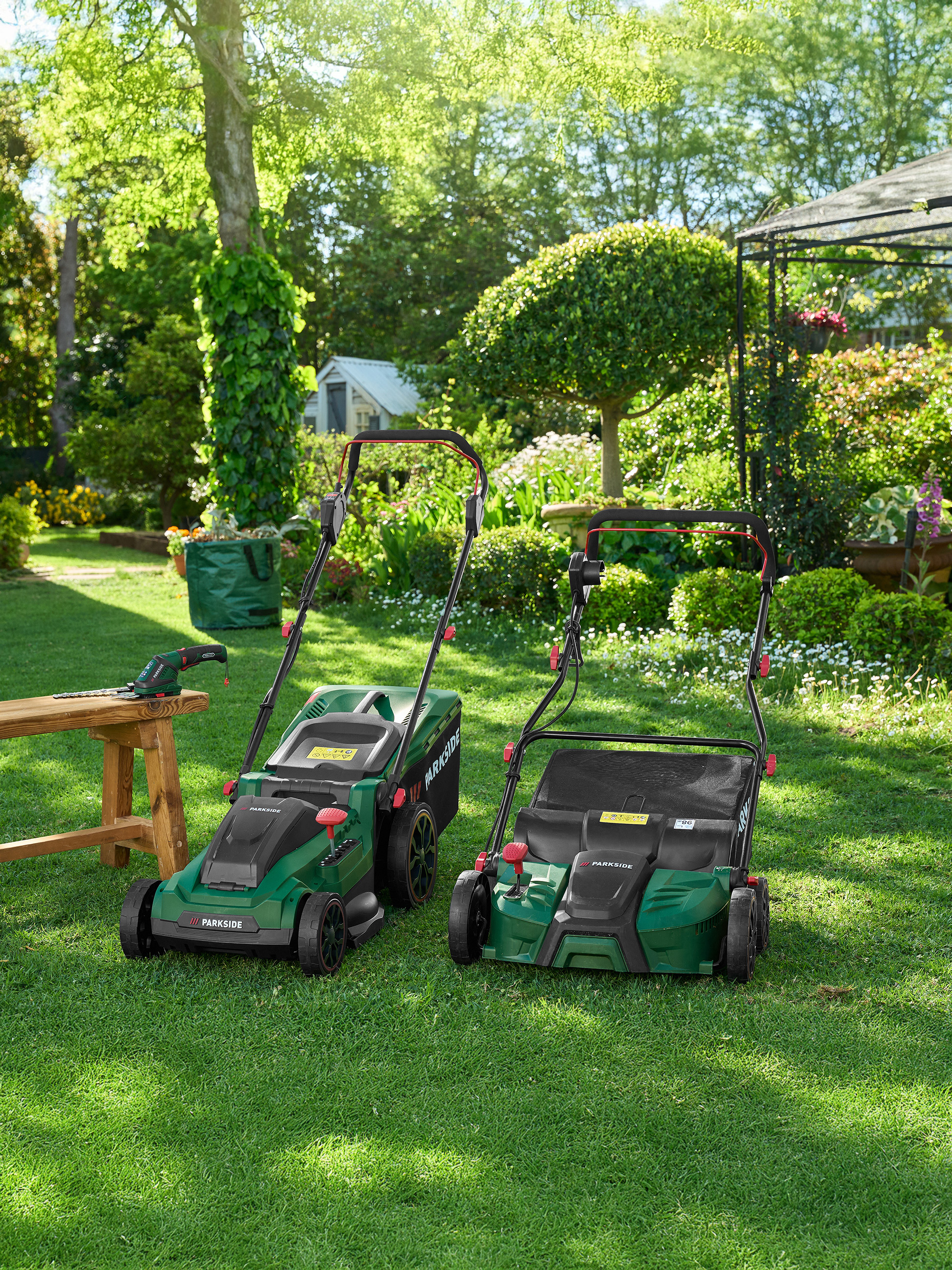 Twee groene Parkside grasmaaiers en tuingereedschap op een gazon in een tuin.