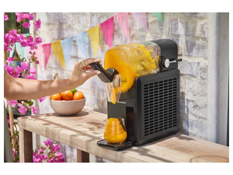 Zwarte slushmachine serveert een sinaasappeldrank in een glas, met feestdecoraties.