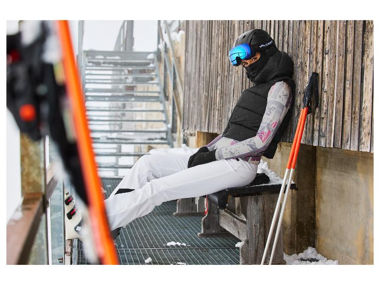 Een vrouw in ski-uitrusting, zwarte bodywarmer en witte broek, zittend op een bankje.