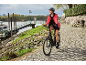 Vrouw met helm en rode jas fietst op een zwarte elektrische fiets op een kasseienpad langs de rivier.
