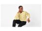 Femme en pull jaune ajouré et pantalon noir, assise sur une chaise blanche.