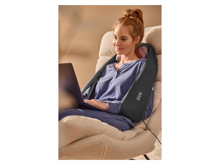 Ontspannen vrouw in een fauteuil gebruikt een nek- en schoudermassager terwijl ze op een laptop werkt.
