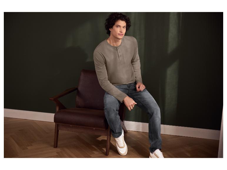 Man in bruin shirt met lange mouwen en blauwe jeans, zittend in een leren fauteuil.