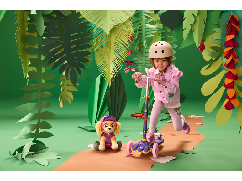 Meisje met helm op een step, naast een knuffelhond, in een jungle-achtige setting.